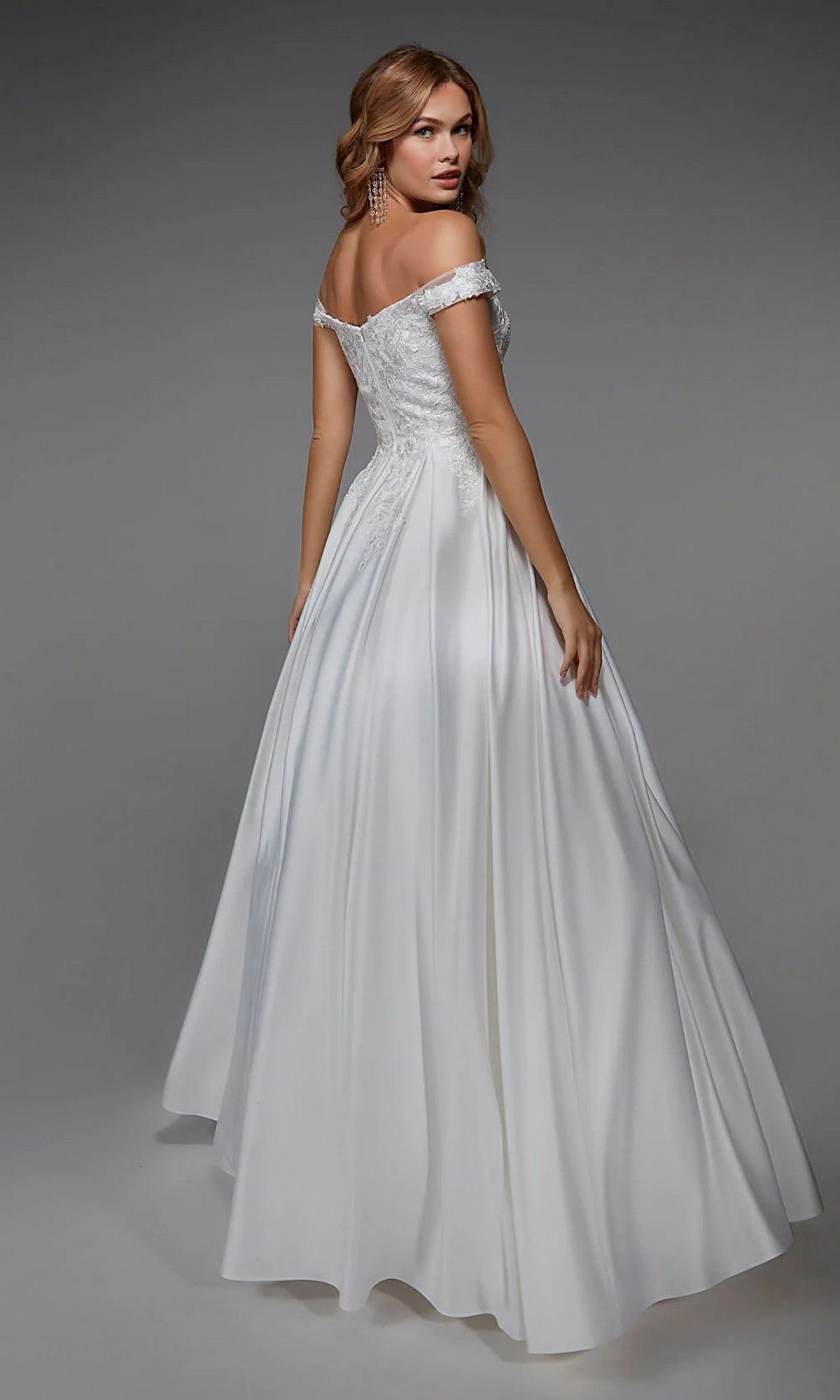 A woman in the Long White Dress: Alyce 7034, featuring a floral embroidered bodice and flowing skirt, poses in an off-the-shoulder A-line wedding gown against a plain background.